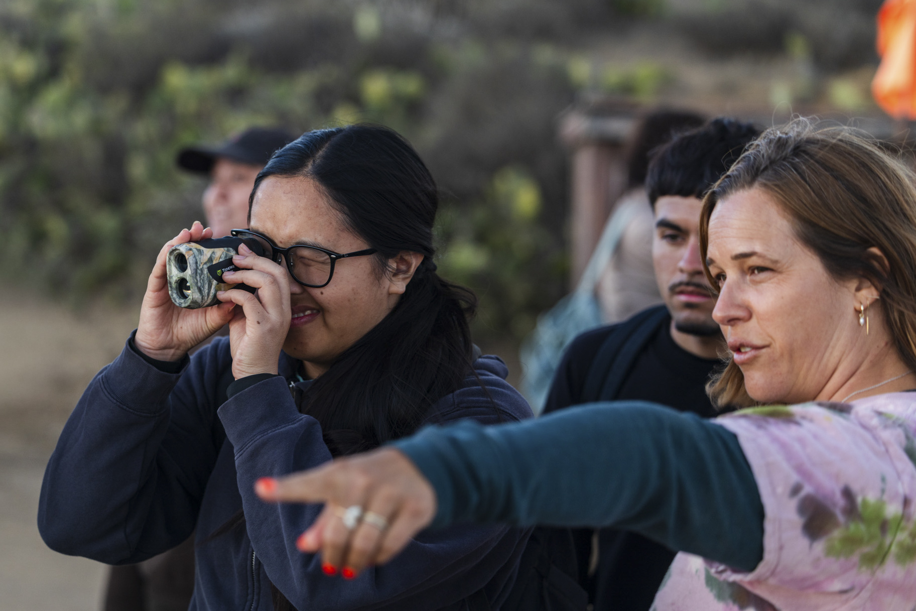 A person looks through a handheld rangefinder while another person points toward the distance